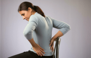  A young woman sitting at an office desk suffering from acute back pain and poor postural alignment. 